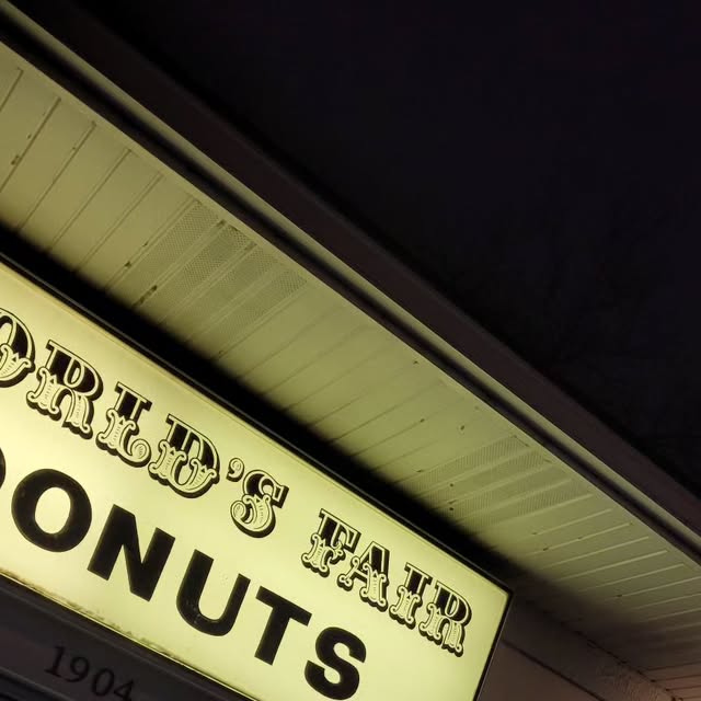 The World's Fair Donuts storefront sign lit up at night, WORLD'S FAIR in ornate lettering above DONUTS, with 1904 beneath
